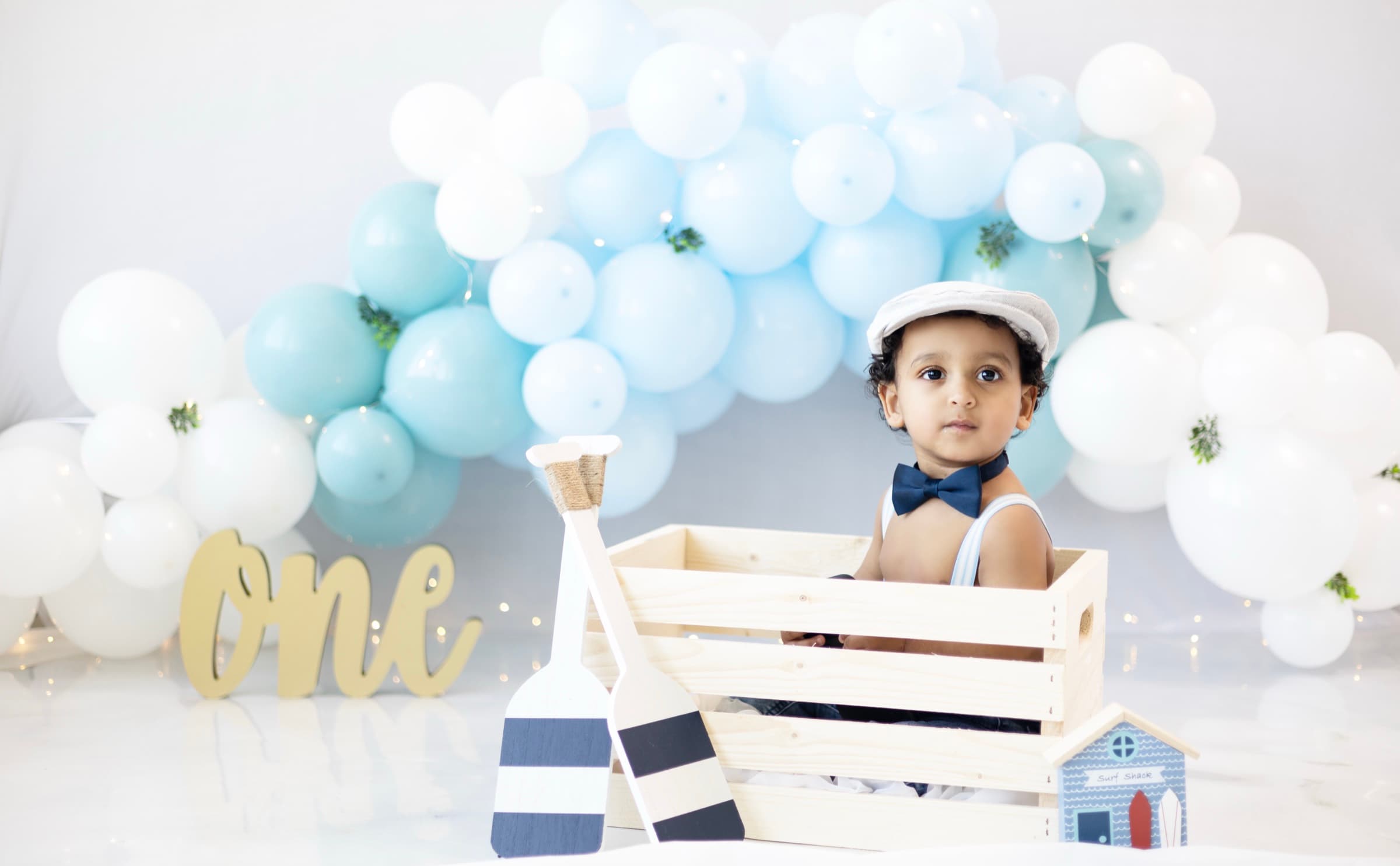 A one-year-old boy in a newsboy cap sits in a wooden crate surrounded by nautical birthday props and blue balloons.