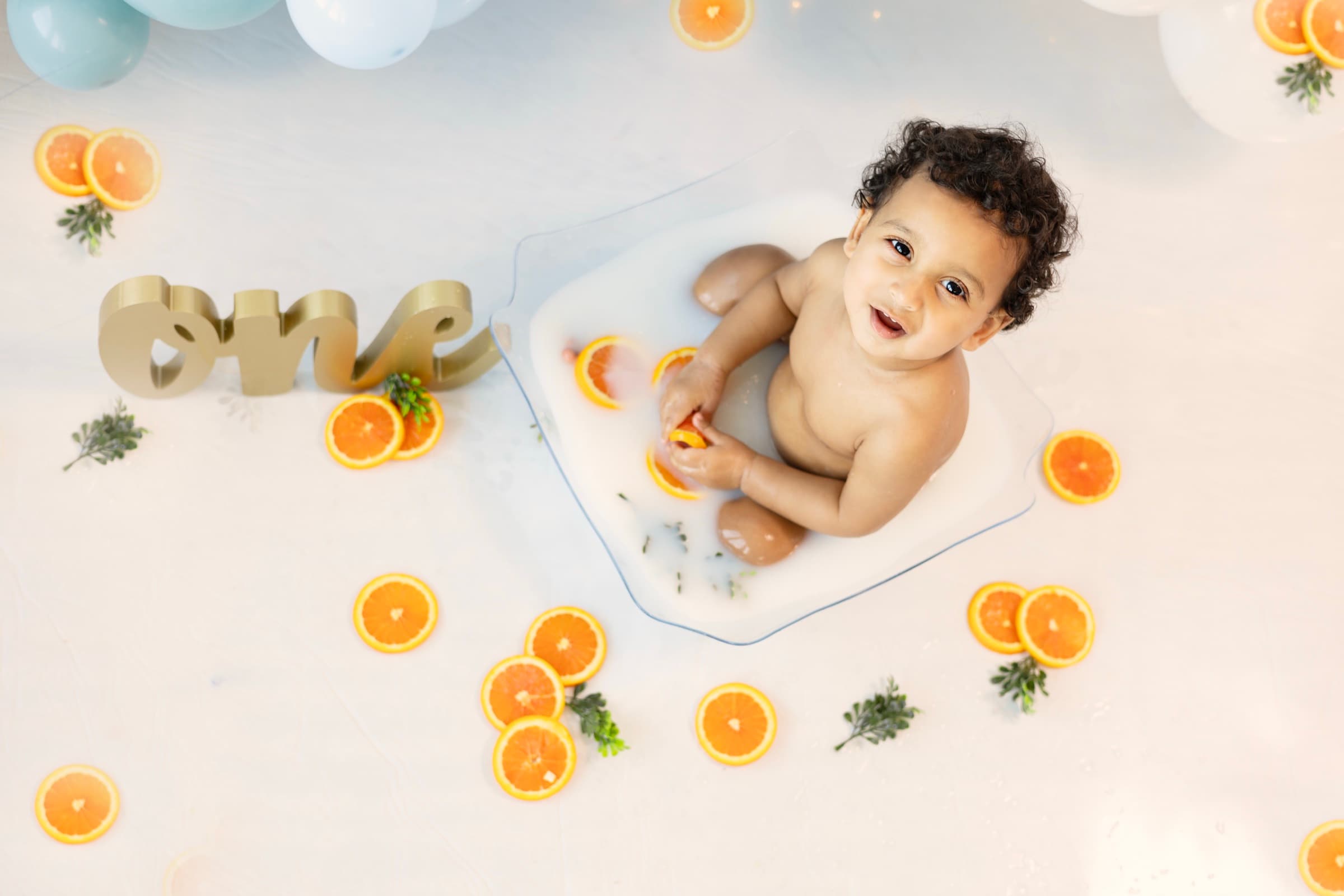 Overhead view of a one-year-old boy laughing in a milk bath scattered with orange slices, surrounded by a blue and white balloon garland.