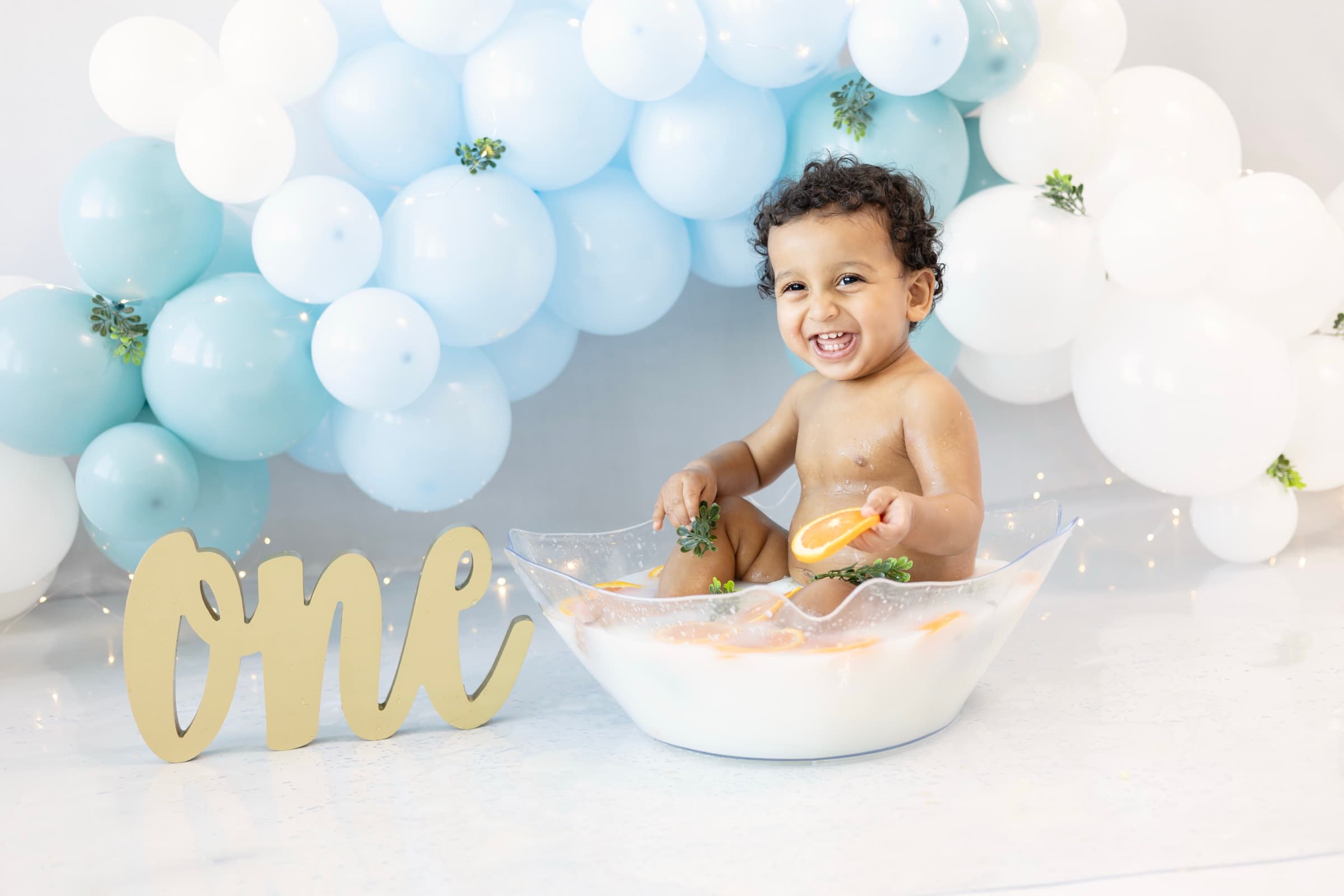 A one-year-old boy laughs joyfully in a milk bath, holding an orange slice, blue balloon garland behind him.