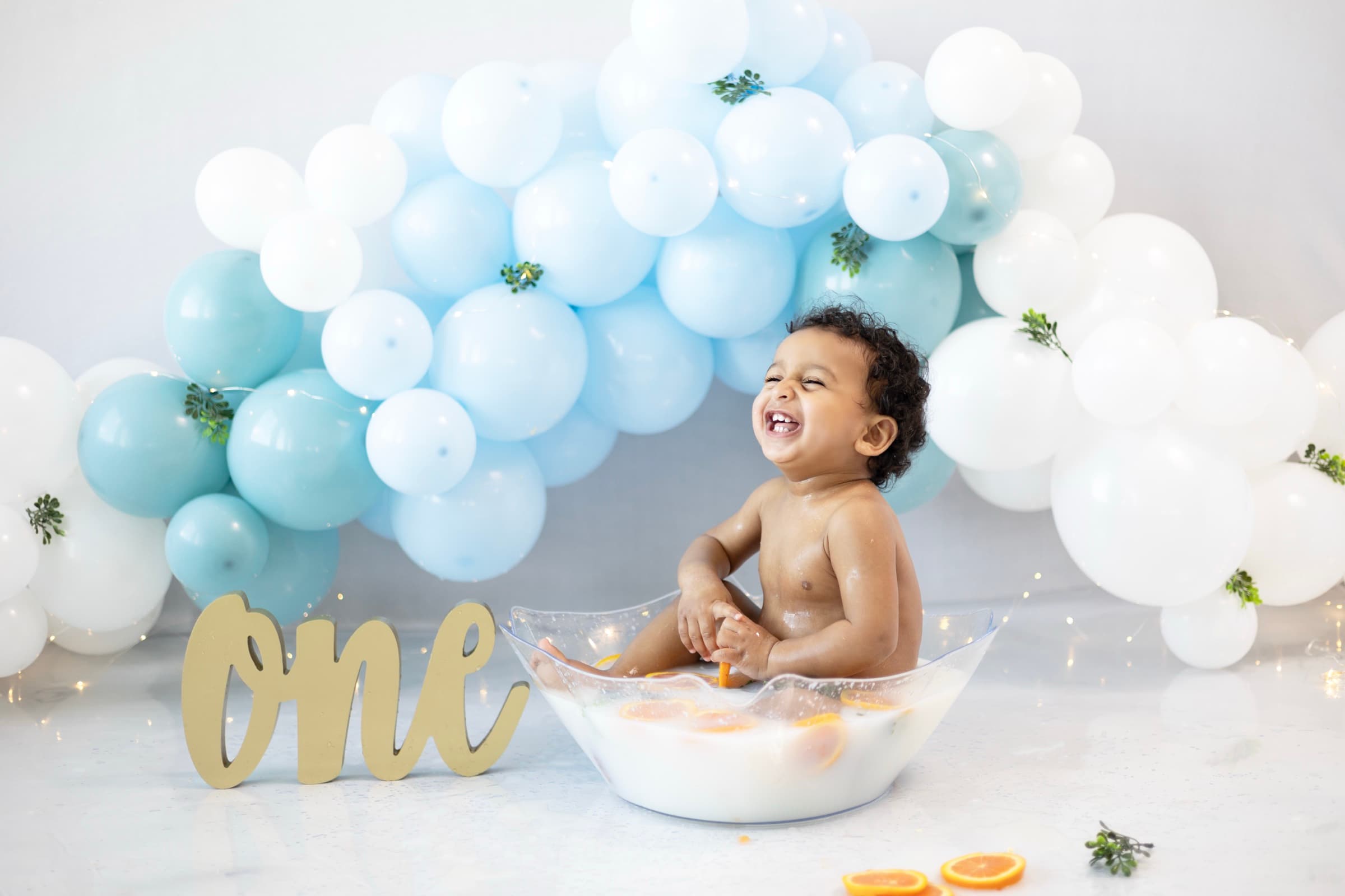 A one-year-old boy laughs with his eyes closed in a milk bath scattered with orange slices.