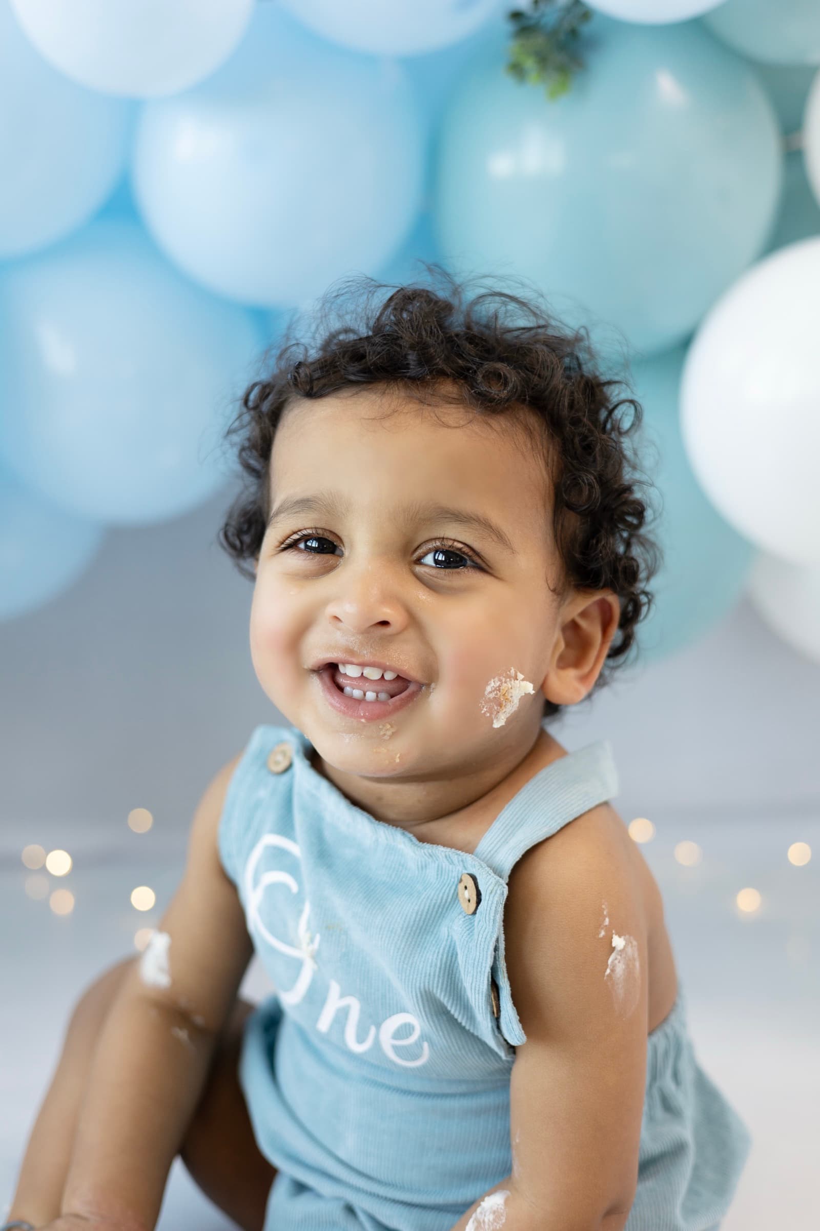 Close-up of a one-year-old boy smiling directly at the camera with birthday cake on his cheek, warm balloon backdrop.