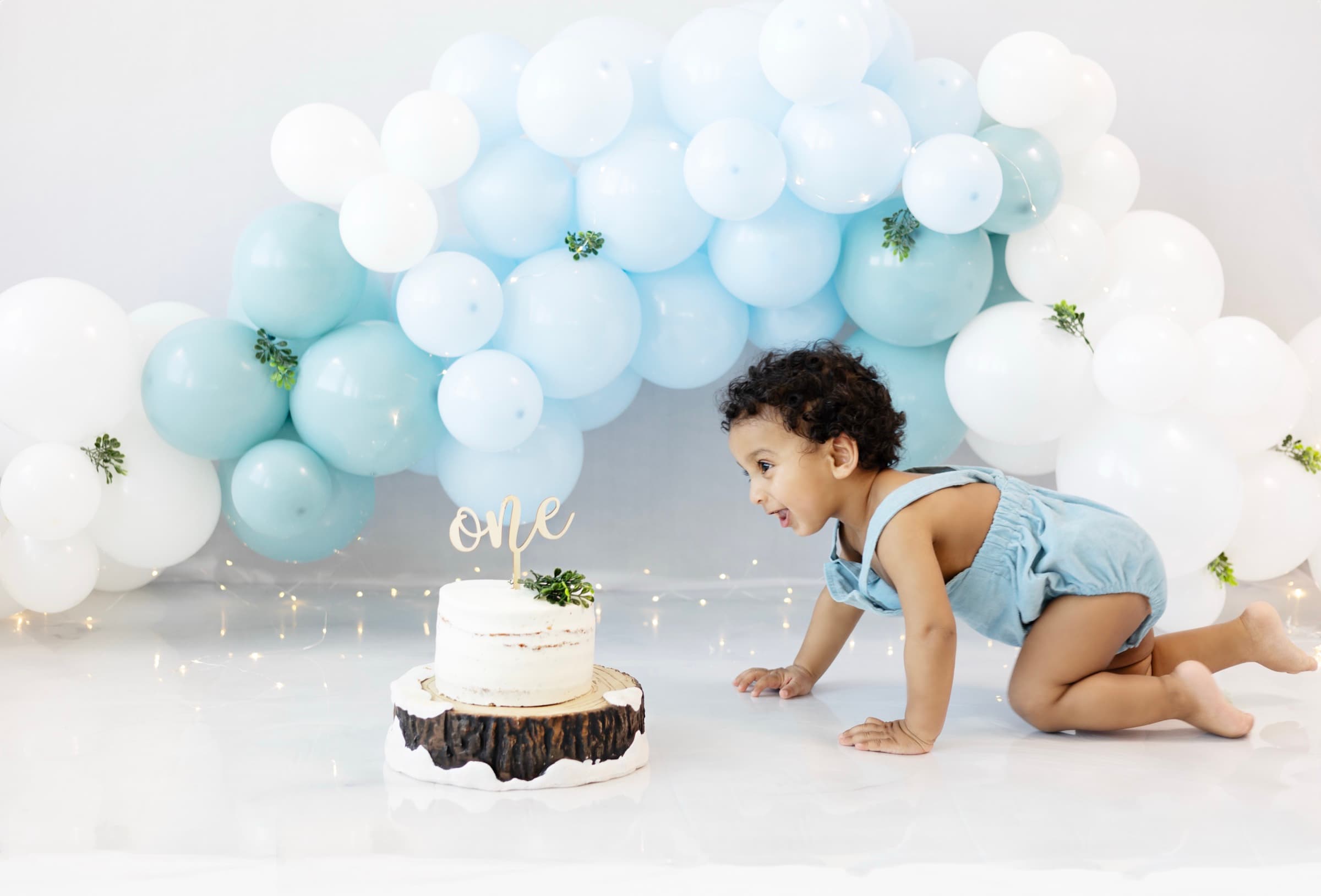 A one-year-old boy in a blue romper crawls eagerly toward his birthday cake, balloon garland overhead.