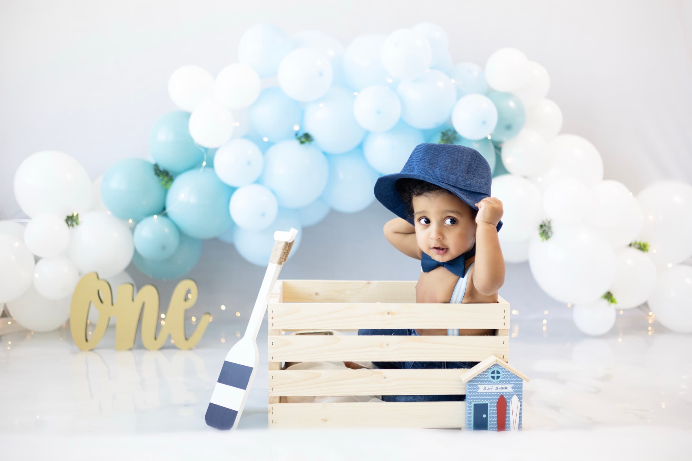 A one-year-old boy in a denim bucket hat sits in a wooden crate with nautical birthday props.