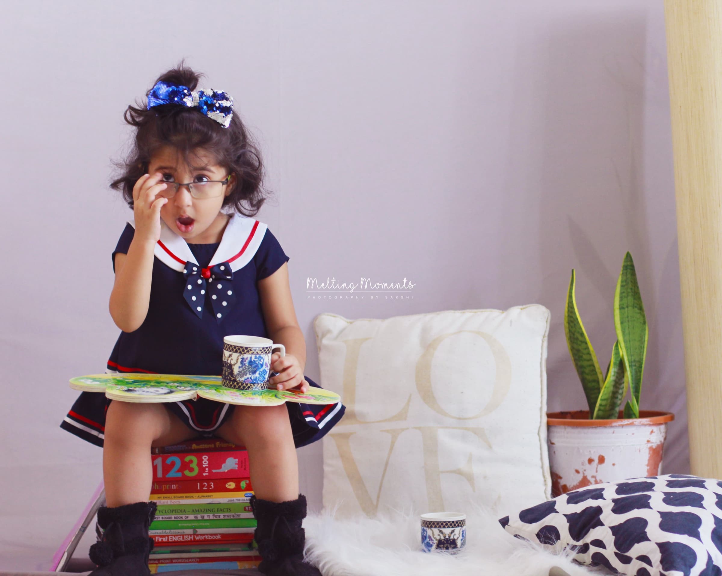 A young girl in a sailor dress and glasses gasps in mock surprise, holding a teacup, seated on a stack of books.