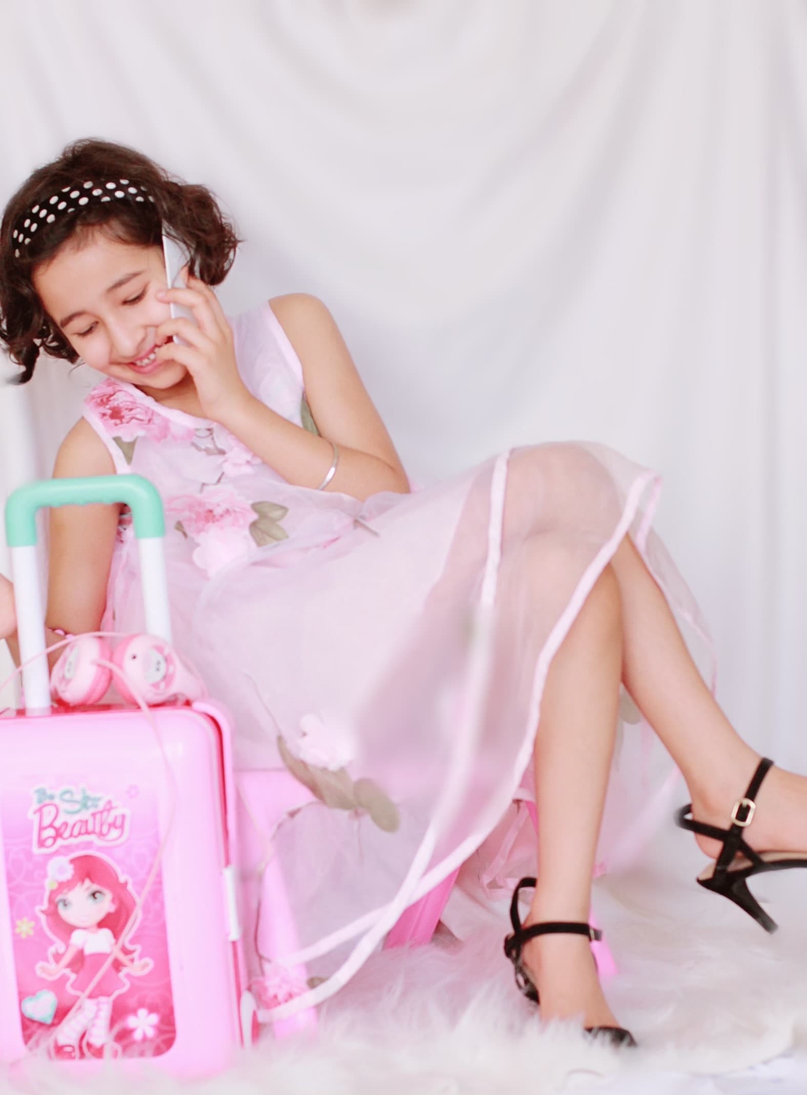 A young girl in a pink dress and heels sits on a pink toy suitcase, chatting on a toy phone.