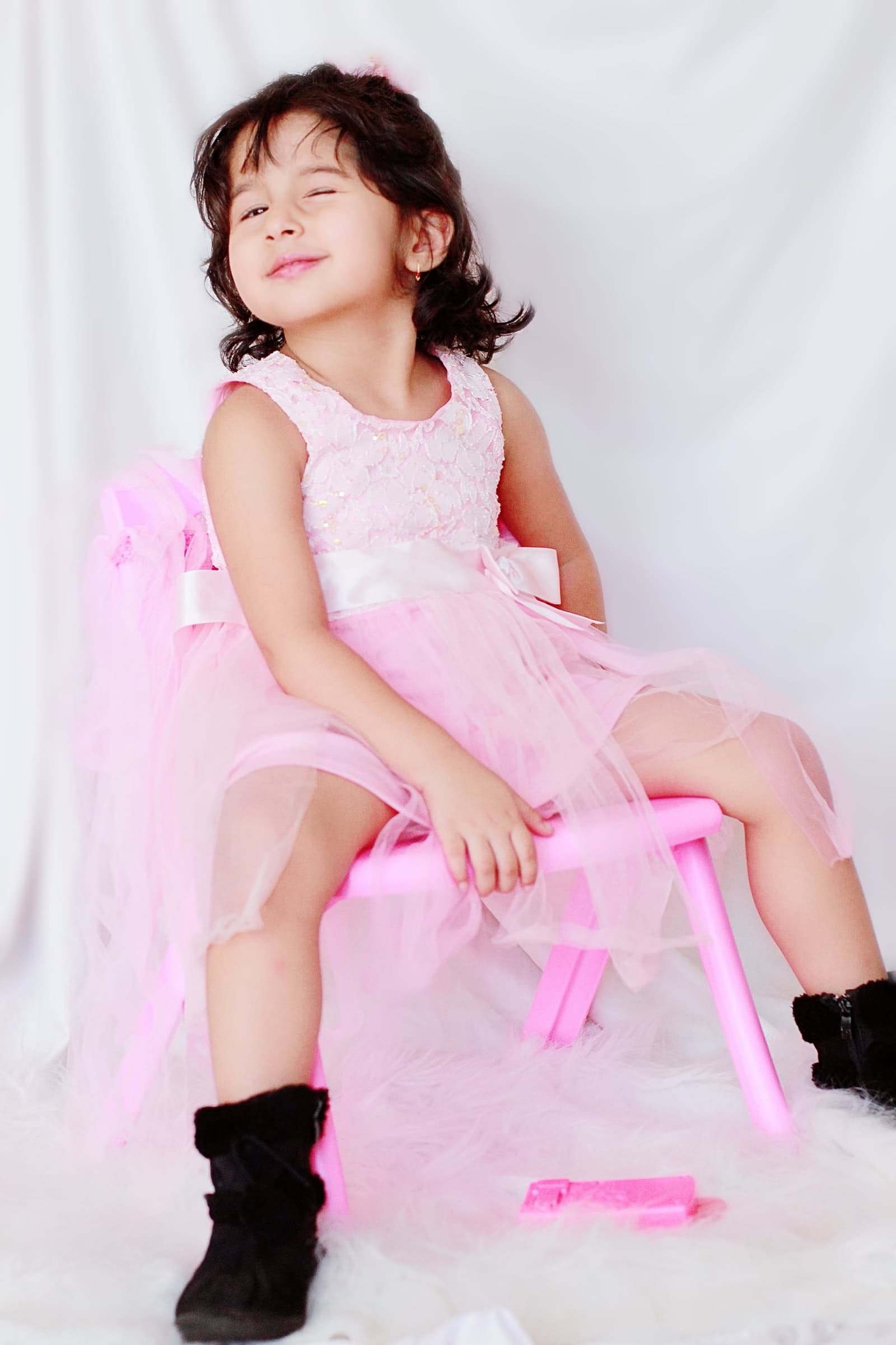A young girl in a pink tulle dress and black boots sits sideways on a chair, chin tilted playfully toward the camera.
