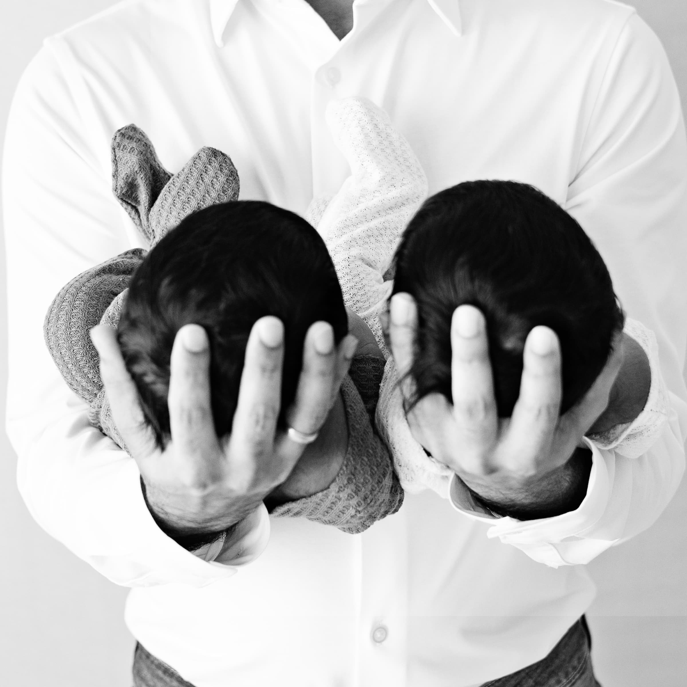 Black-and-white close-up of a father's hands cradling the heads of his newborn twins against his chest.