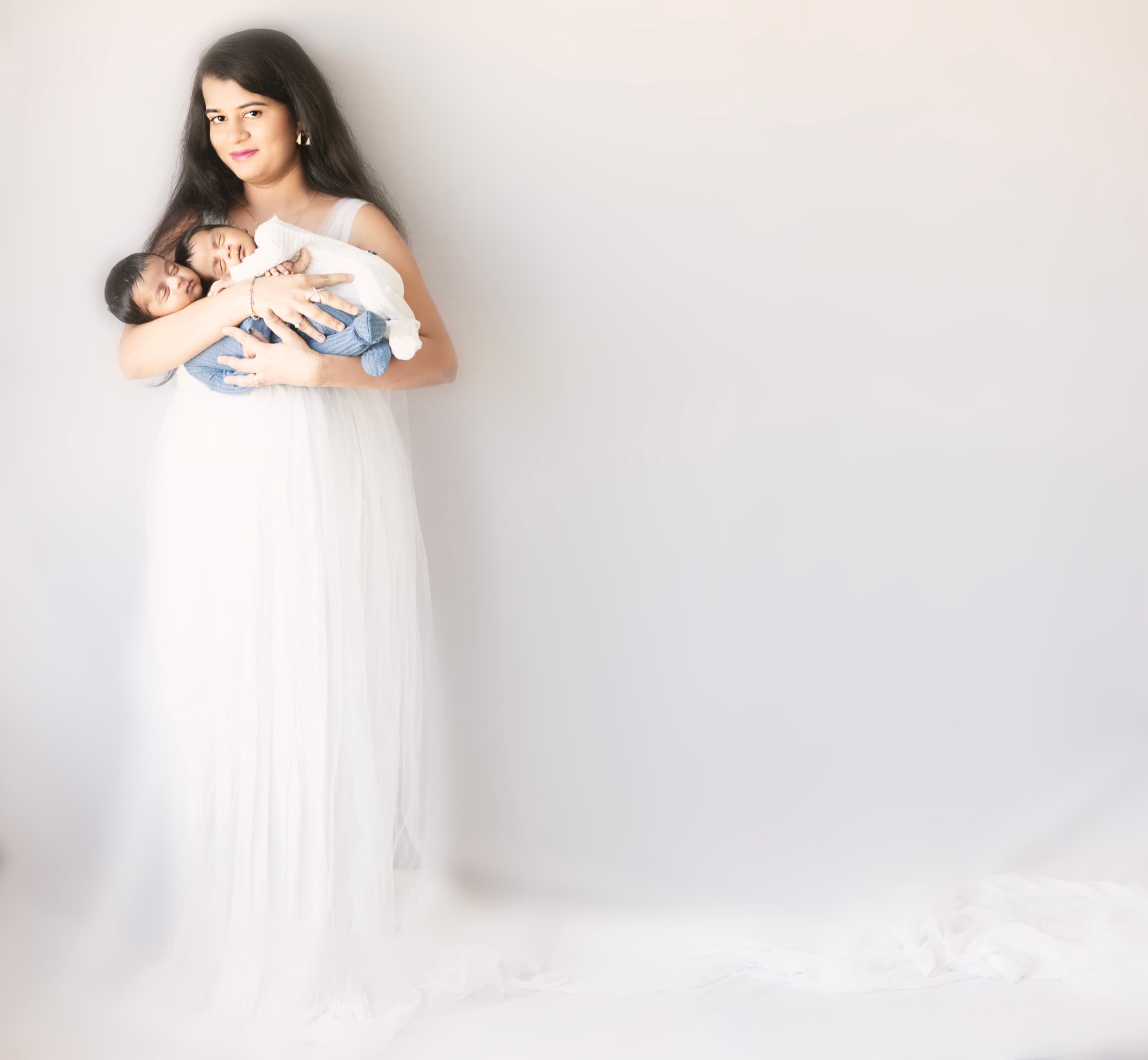 A mother in a flowing white dress holds her newborn twins against her shoulder, soft ethereal backlight.