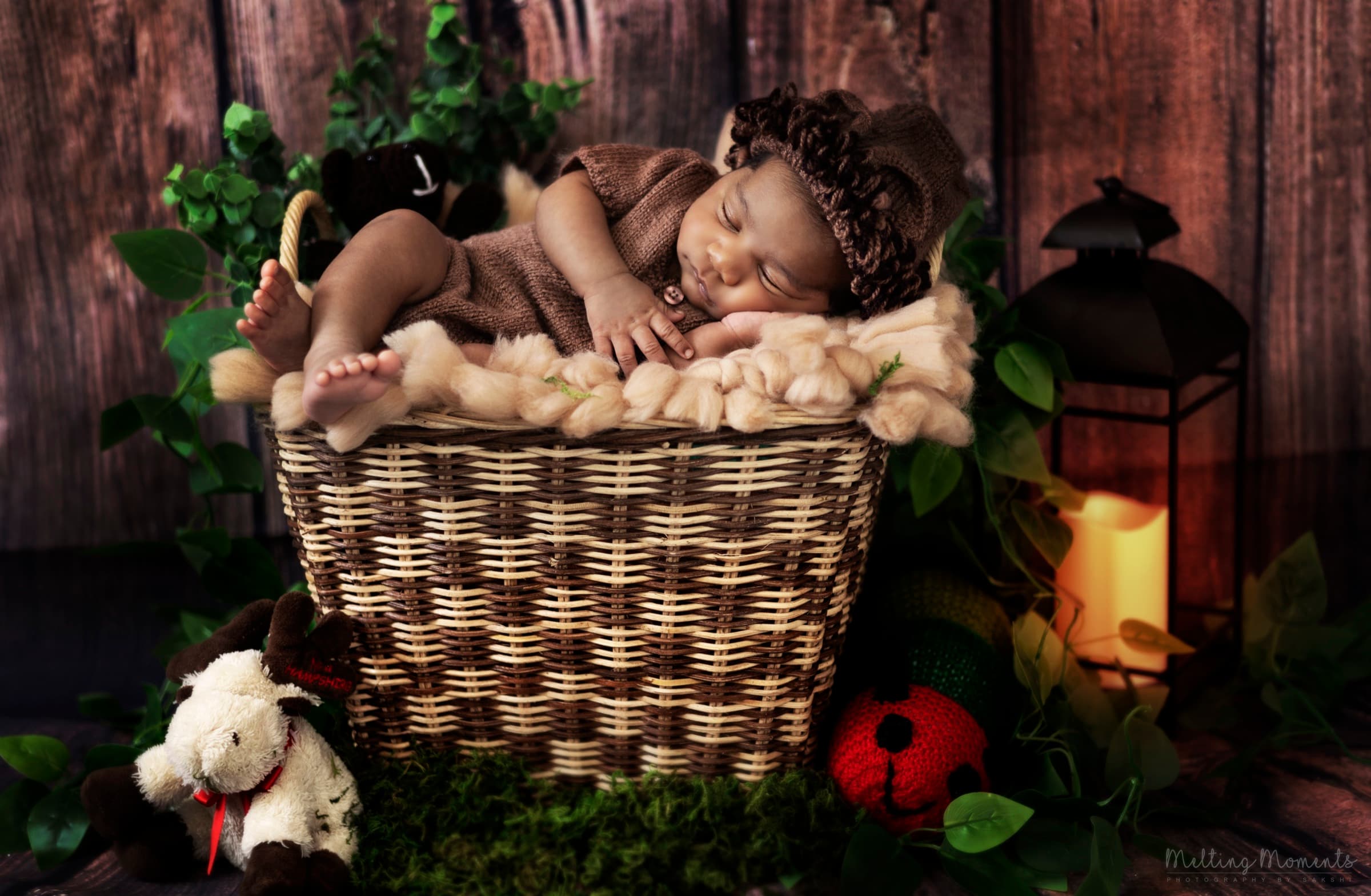 A newborn baby asleep in a woven basket wearing a knit bonnet, surrounded by greenery and a lantern.