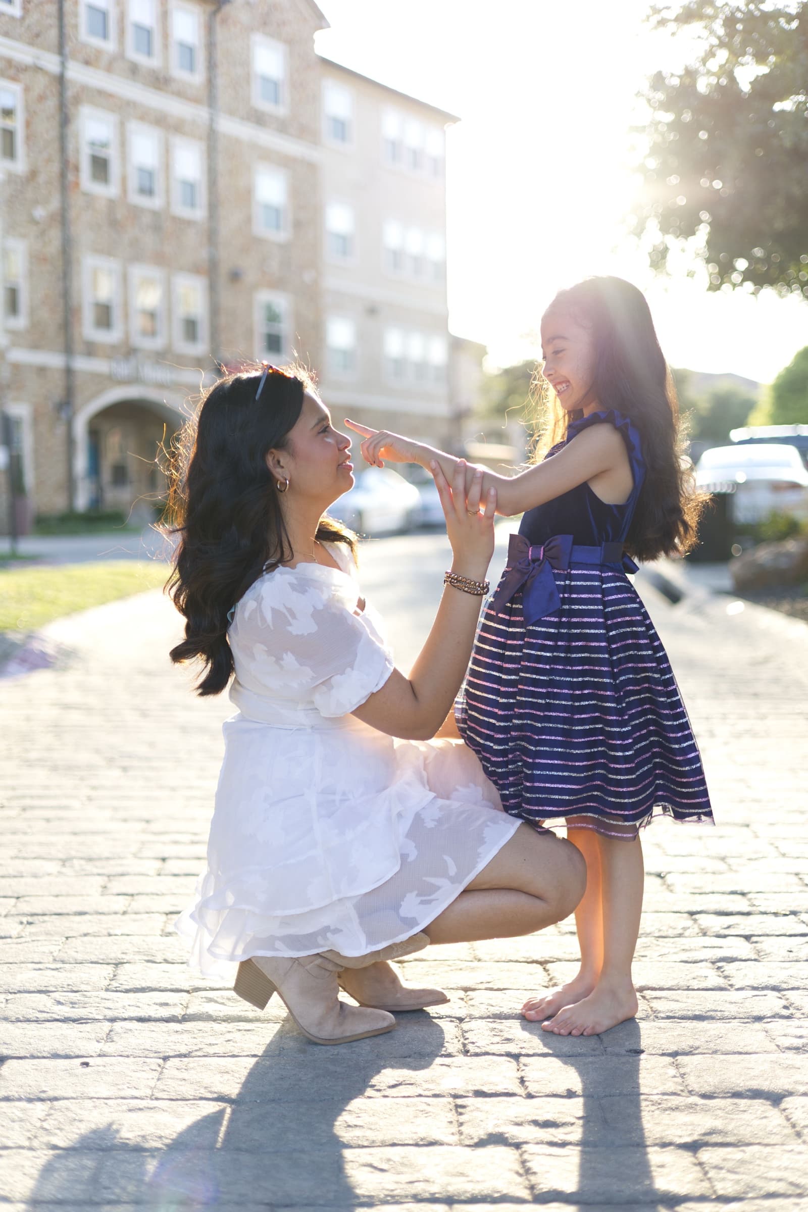 A mother crouches down, playfully touching her young daughter's nose, both laughing in backlit golden light.