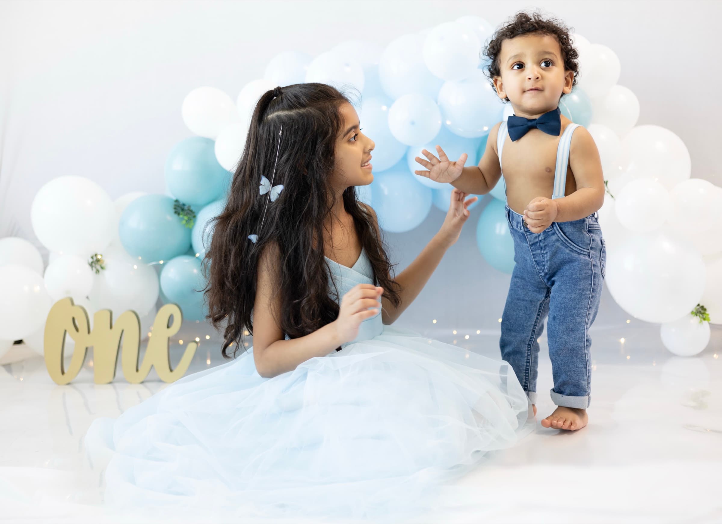 A young girl in a pale blue tulle dress reaches out to her baby brother, who wears suspenders and a bow tie, at a birthday balloon backdrop.