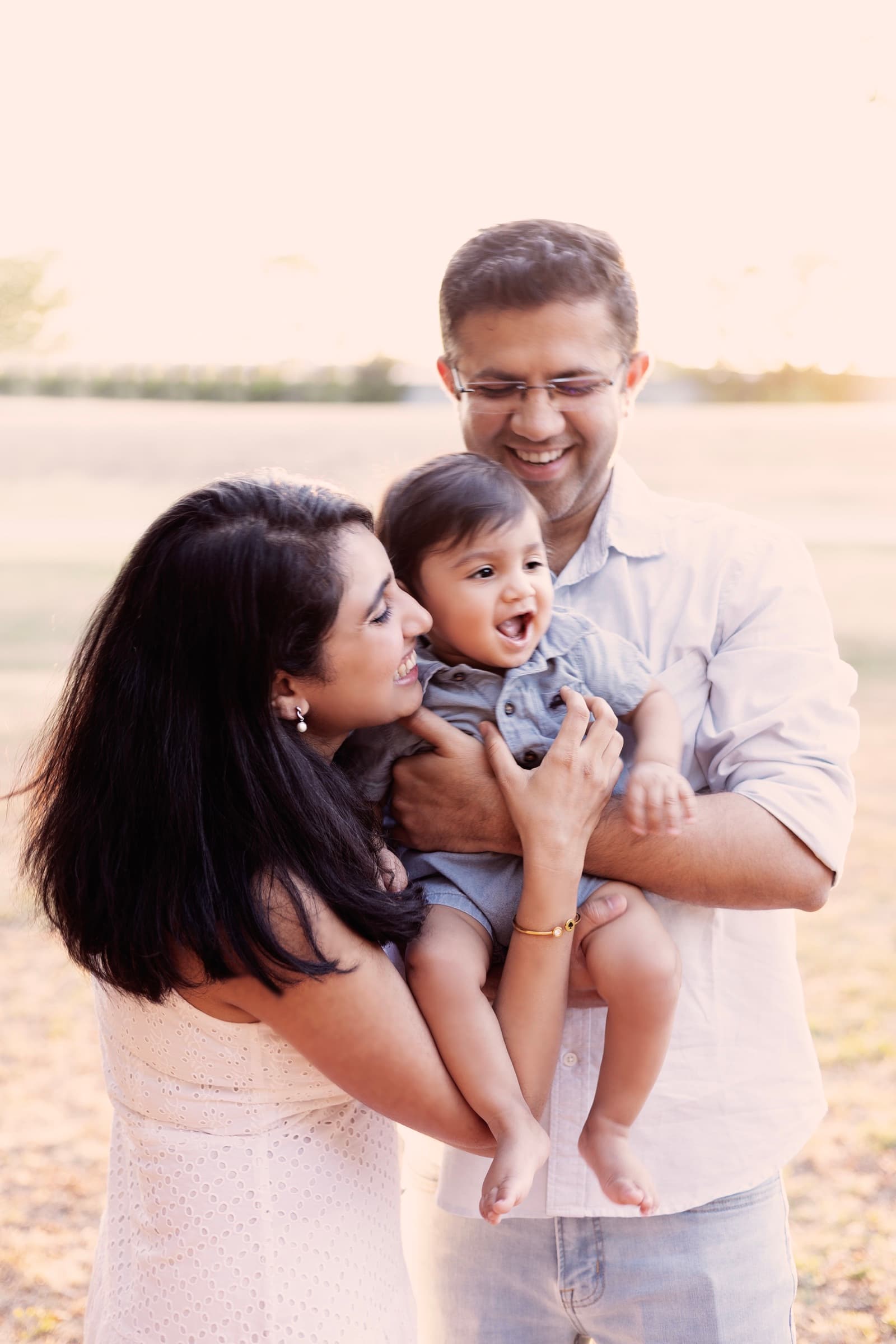 A mother, father, and toddler son laughing together in an open field at golden hour.