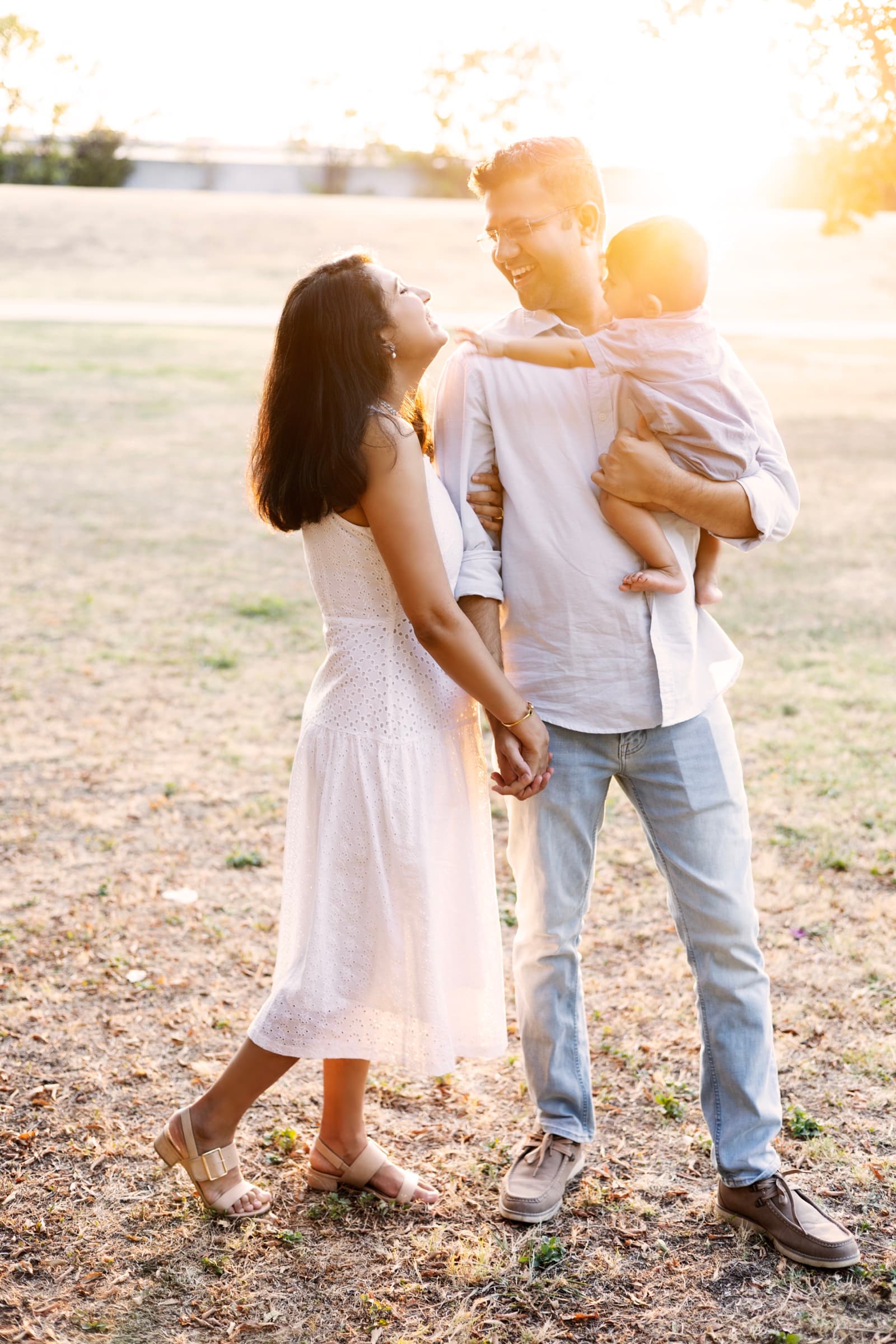 A mother and father laugh with their toddler son, held in his father's arms, backlit by warm evening sun.