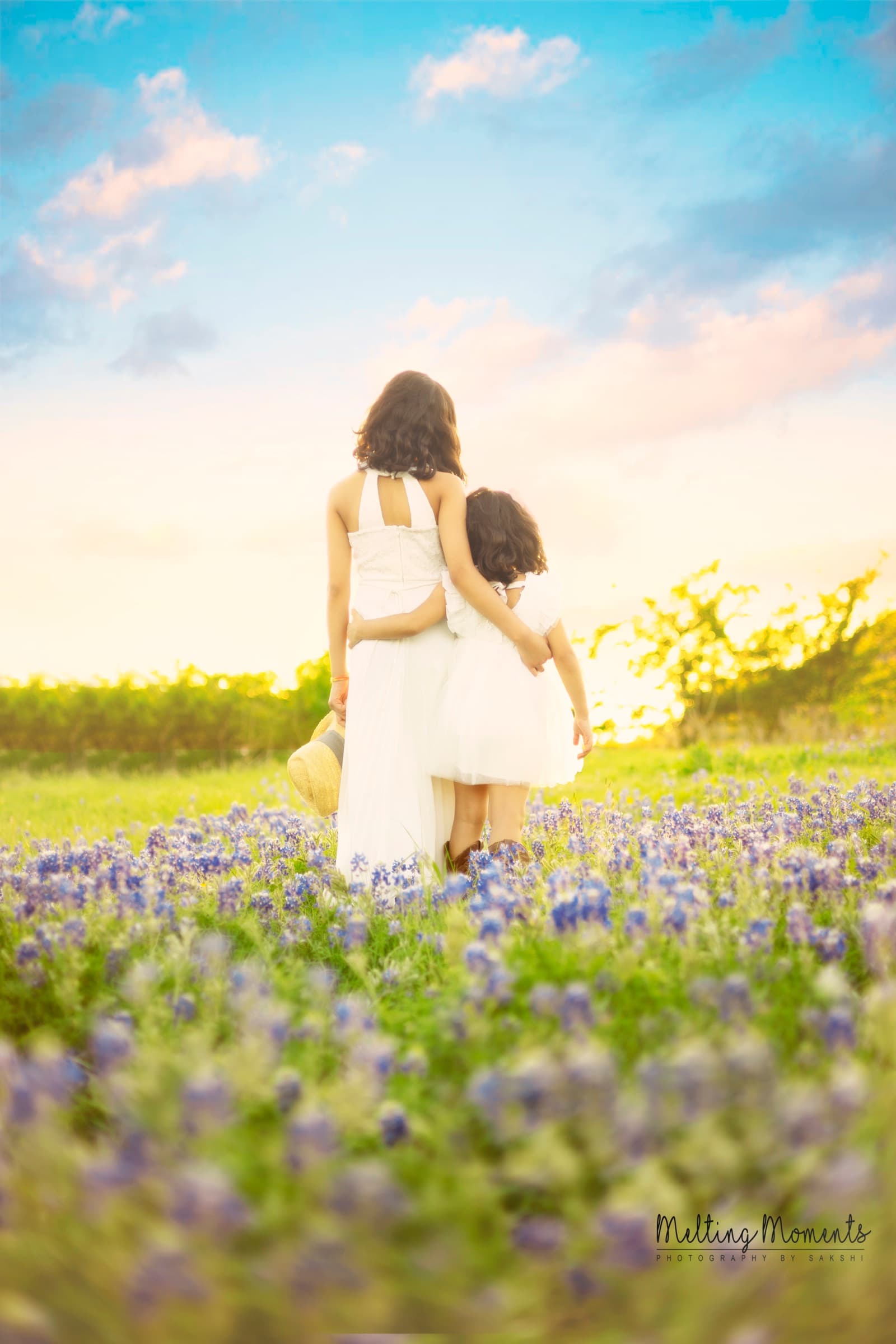 A mother and daughter, both in white dresses, walk hand in hand through a Texas bluebonnet field at sunset.