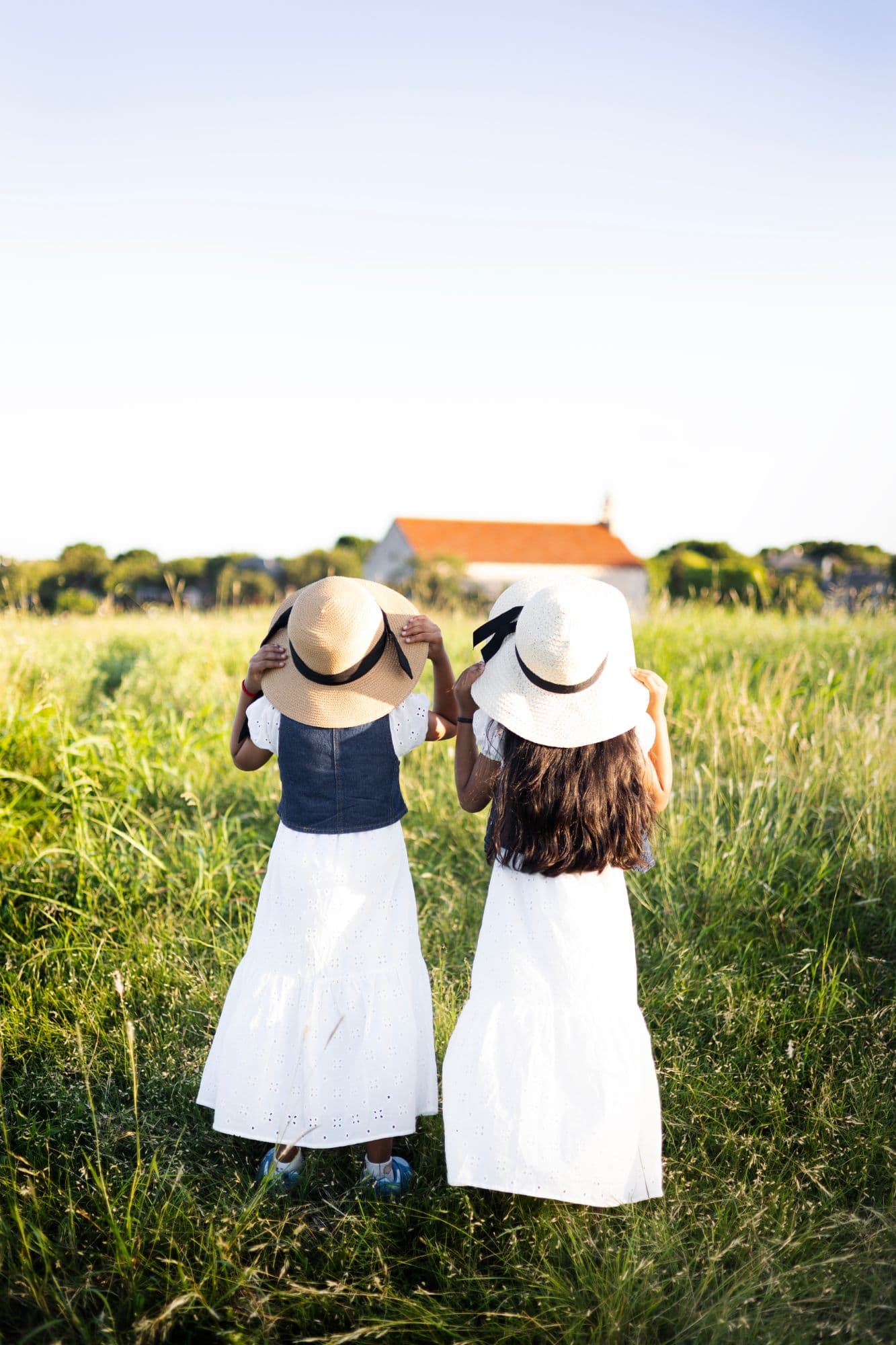 Two young girls in white eyelet dresses and wide-brimmed straw hats, seen from behind, standing in a tall green meadow in golden evening light with an orange-roofed building in the distance.