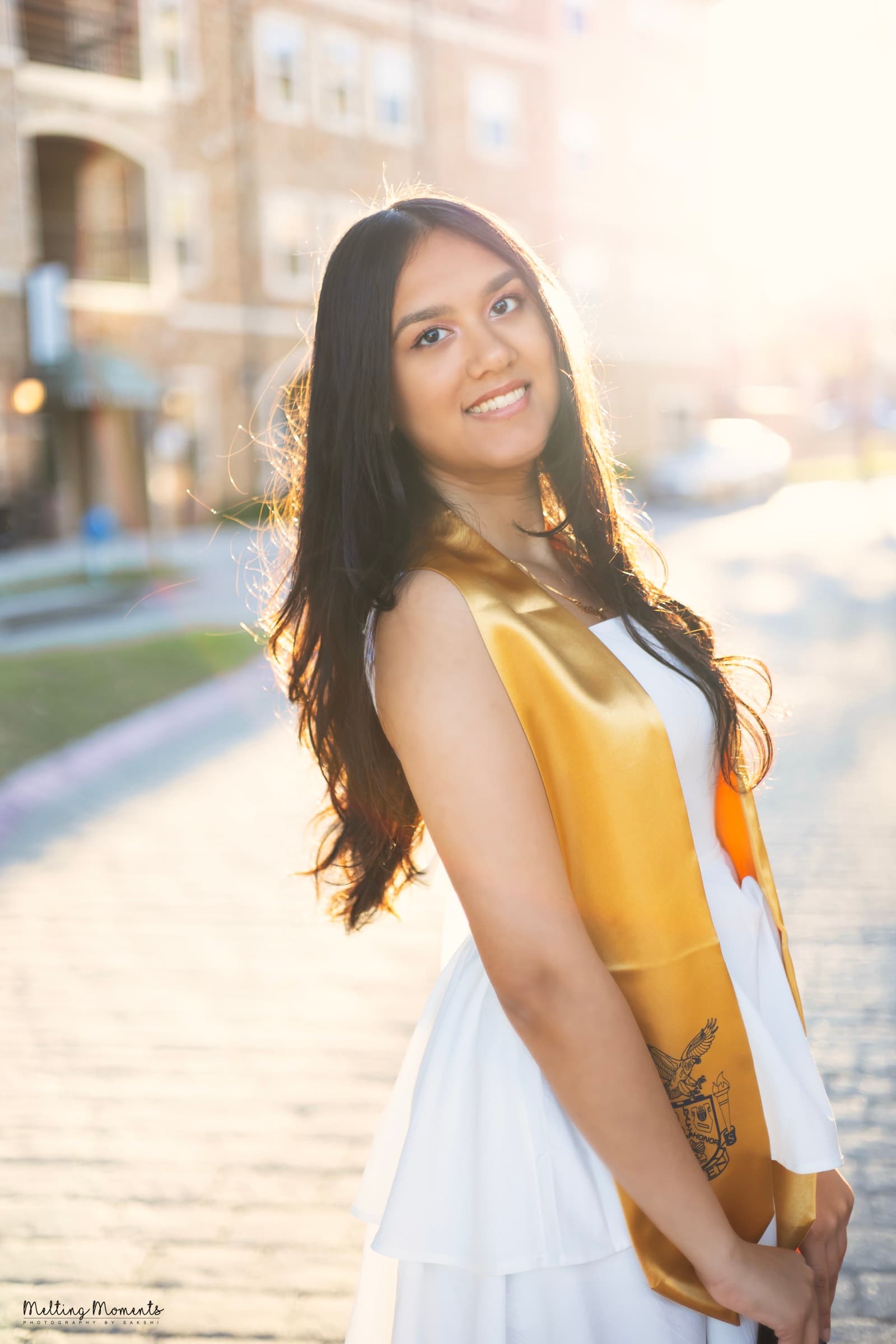 A graduate in a white dress and gold honor sash smiles over her shoulder, backlit by golden hour sun.