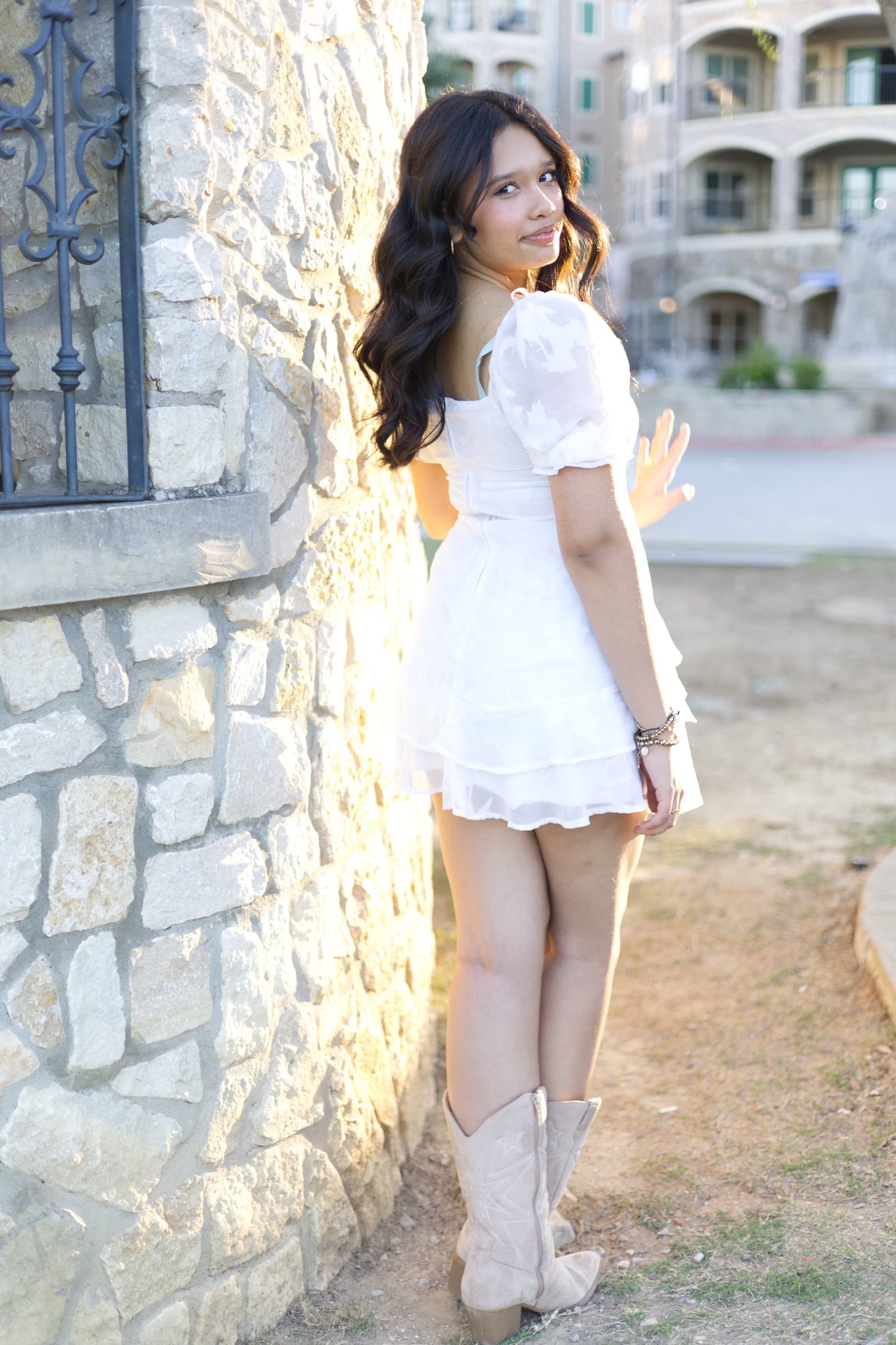 A young woman in a white dress and cowboy boots leans against a stone wall, glancing back over her shoulder in warm evening light.