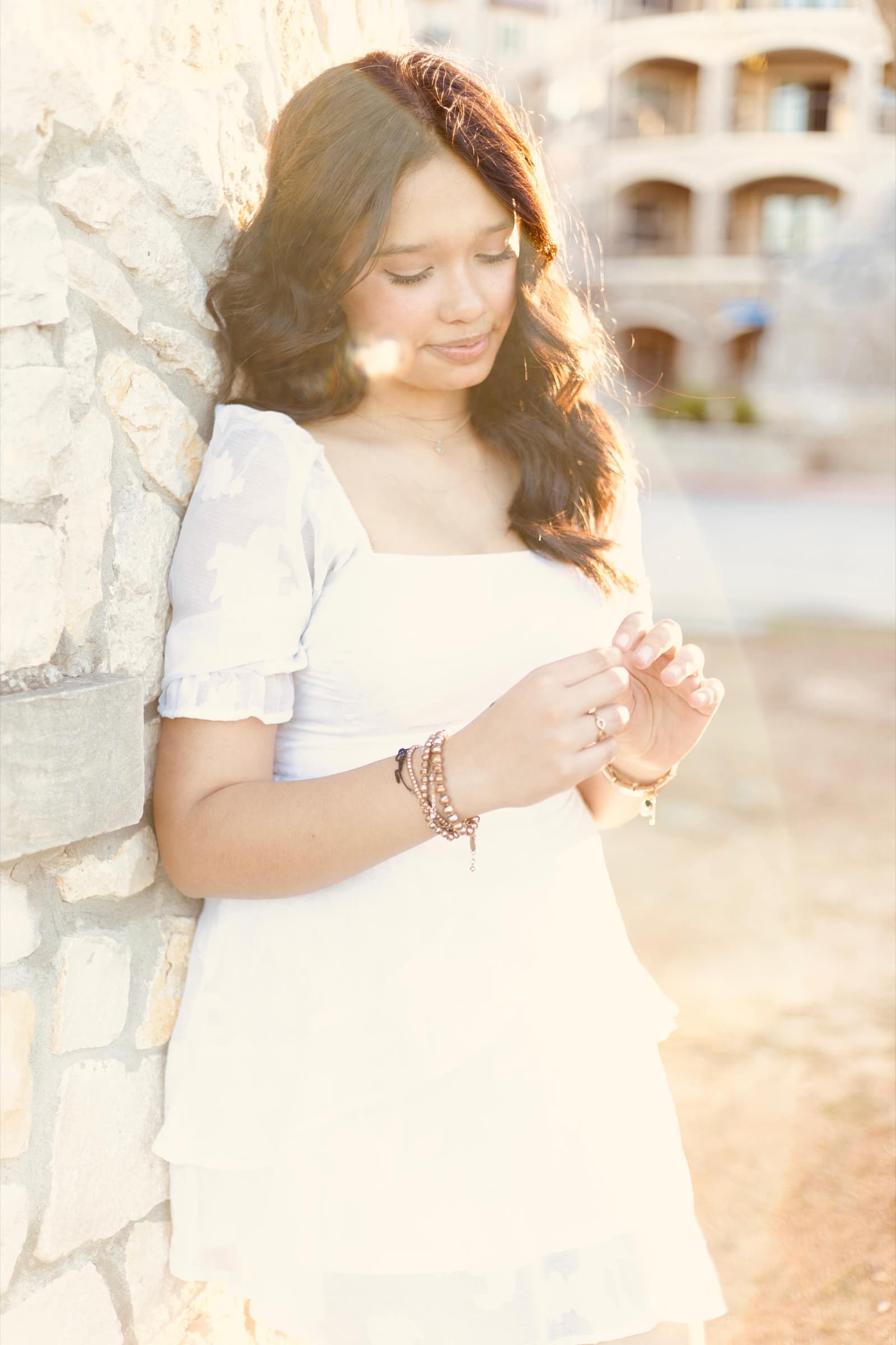 A young woman in a white dress leans against a stone wall, hair caught by the wind, a building softly blurred behind her.