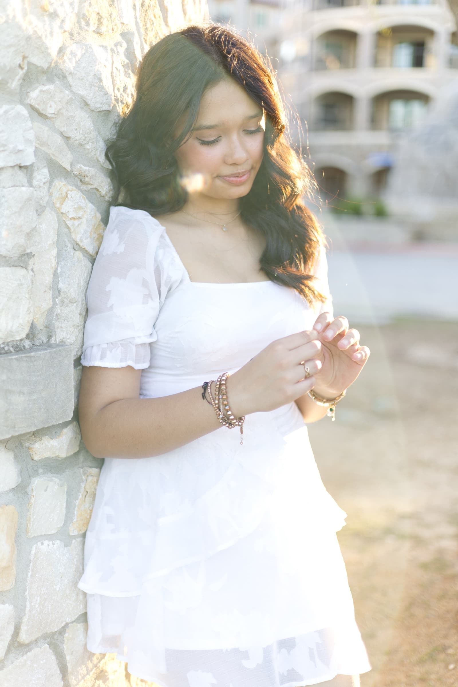 A young woman in a white dress leans against a sunlit stone wall, looking down at her bracelet, soft lens flare across the frame.