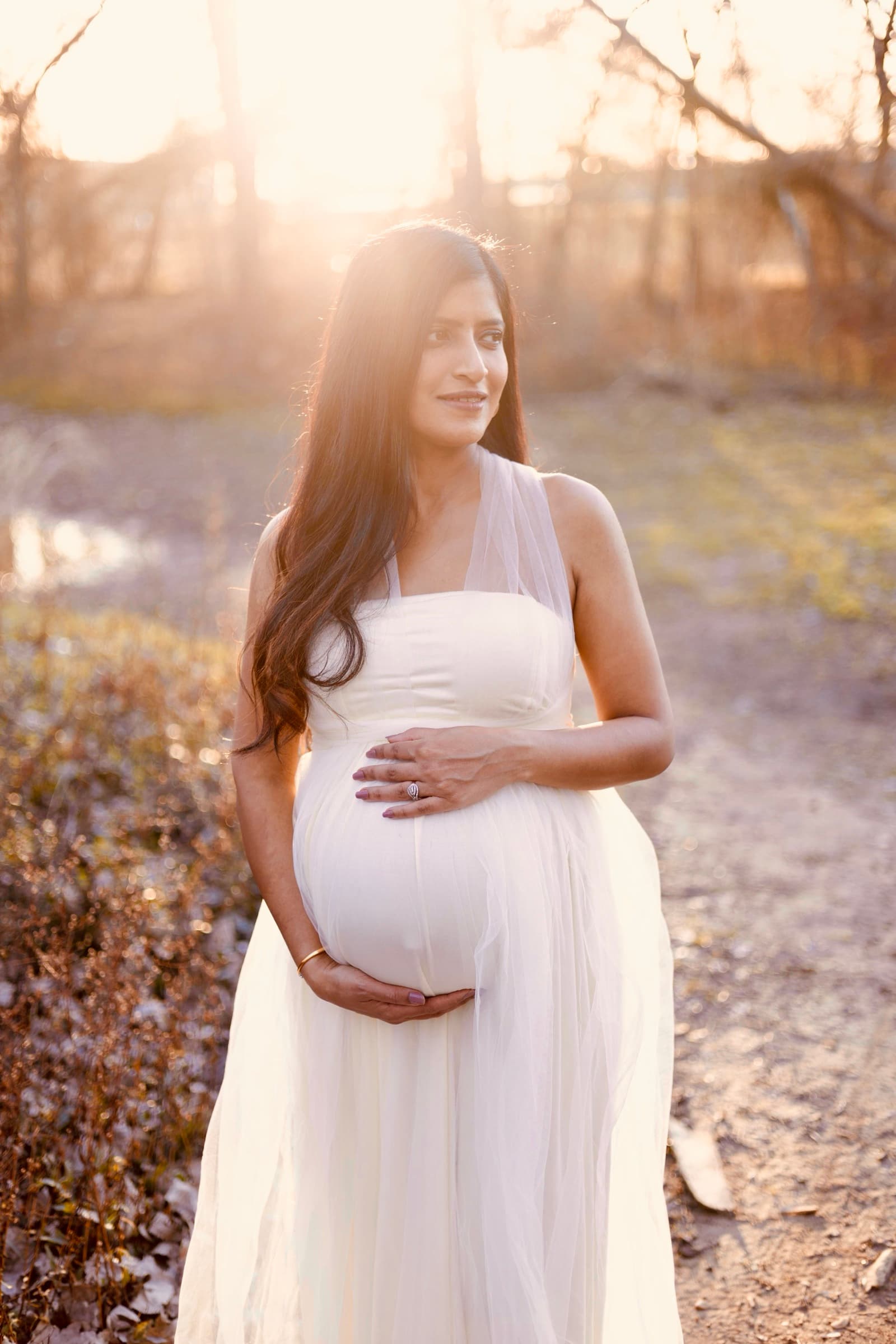 A pregnant woman in a white dress stands near a pond at golden hour, one hand resting on her belly, soft light behind her.