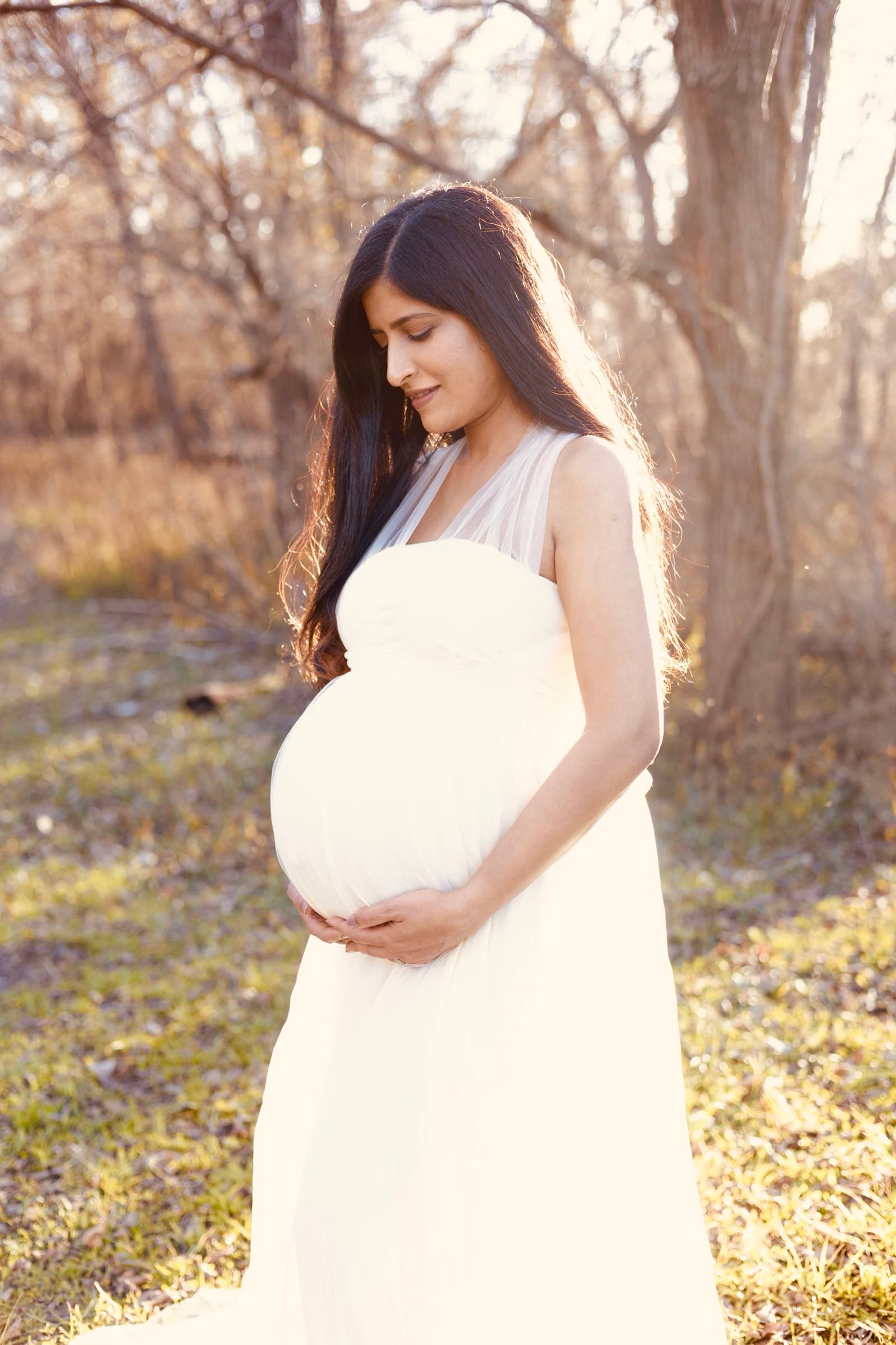 A pregnant woman in a white dress stands in profile, looking down tenderly at her belly, warm backlight through the trees.