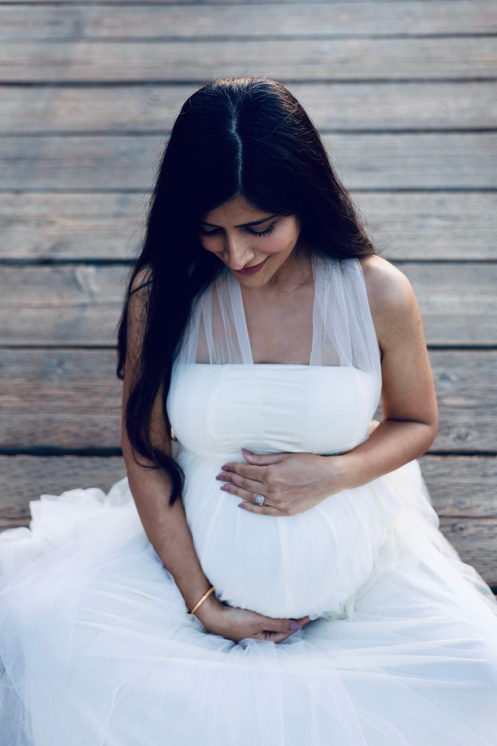 A pregnant woman in a white dress sits on a wooden dock, holding her belly, cooler dusk light and a weathered wood backdrop.