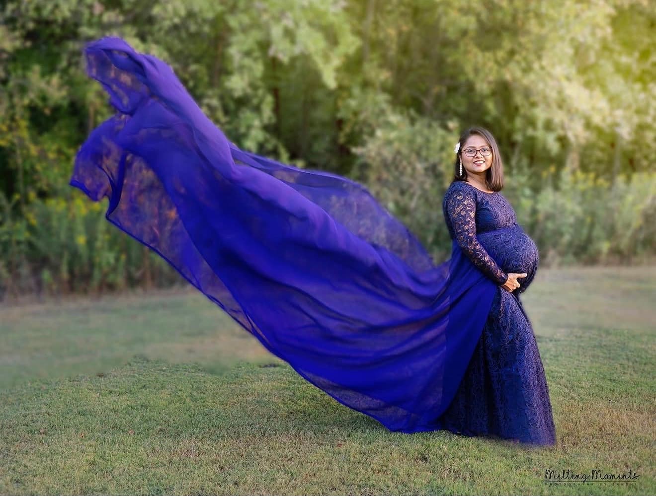 A pregnant woman in a royal blue lace gown smiles at the camera as the dress's long chiffon train catches the wind across a grassy field.