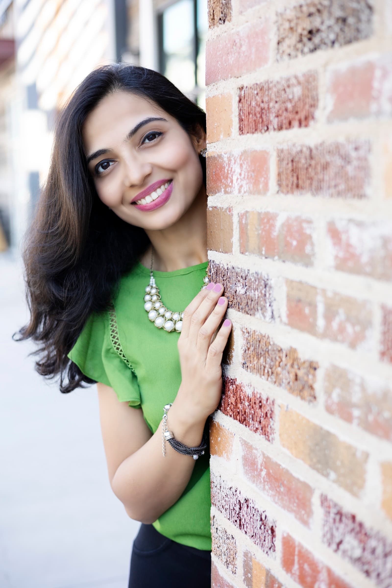 A woman in a green top smiles warmly from behind a brick wall corner, sunlight on her face.