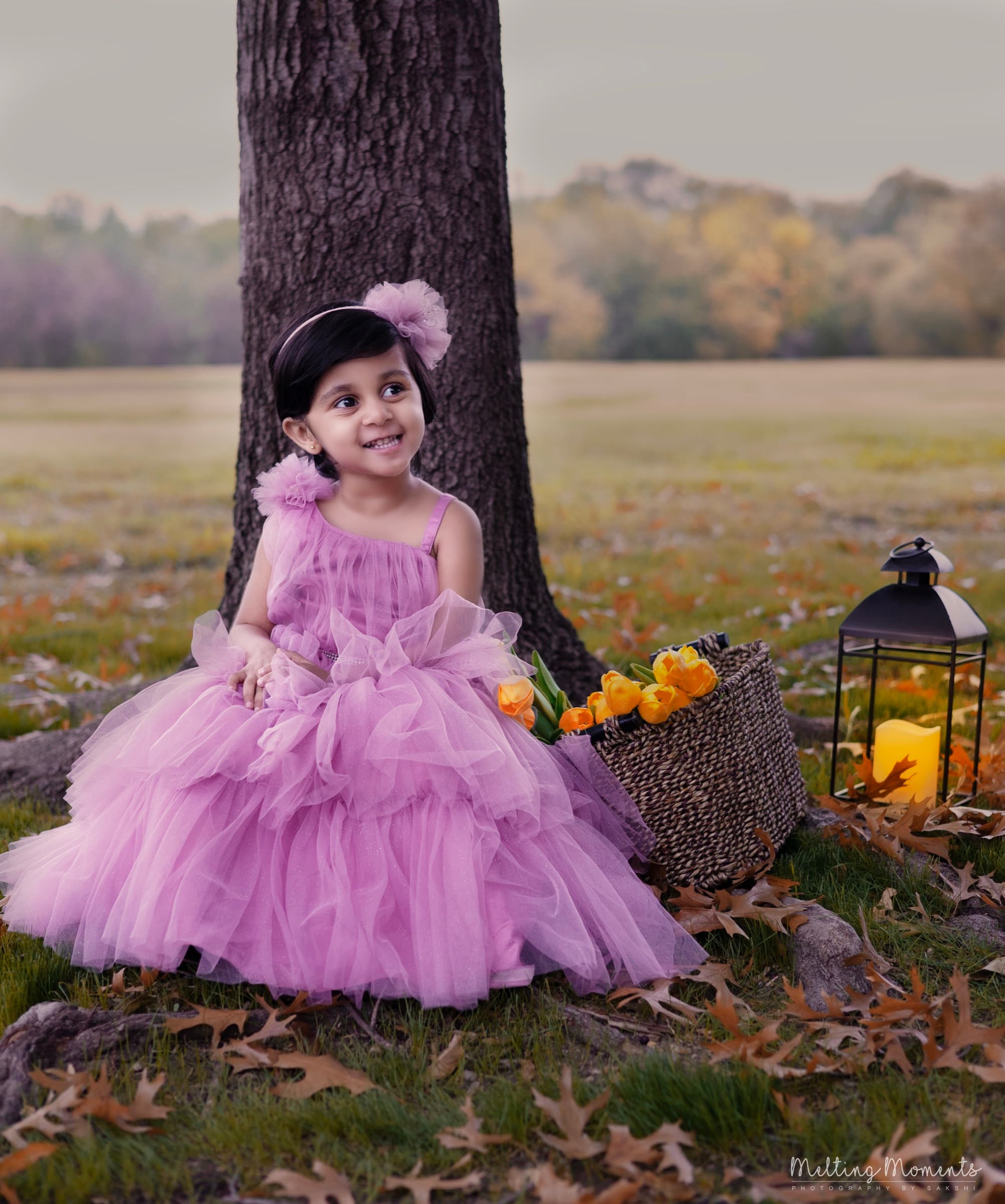 A young girl in a lavender tulle dress sits at the base of a tree in a golden autumn field, surrounded by yellow tulips and a lantern.