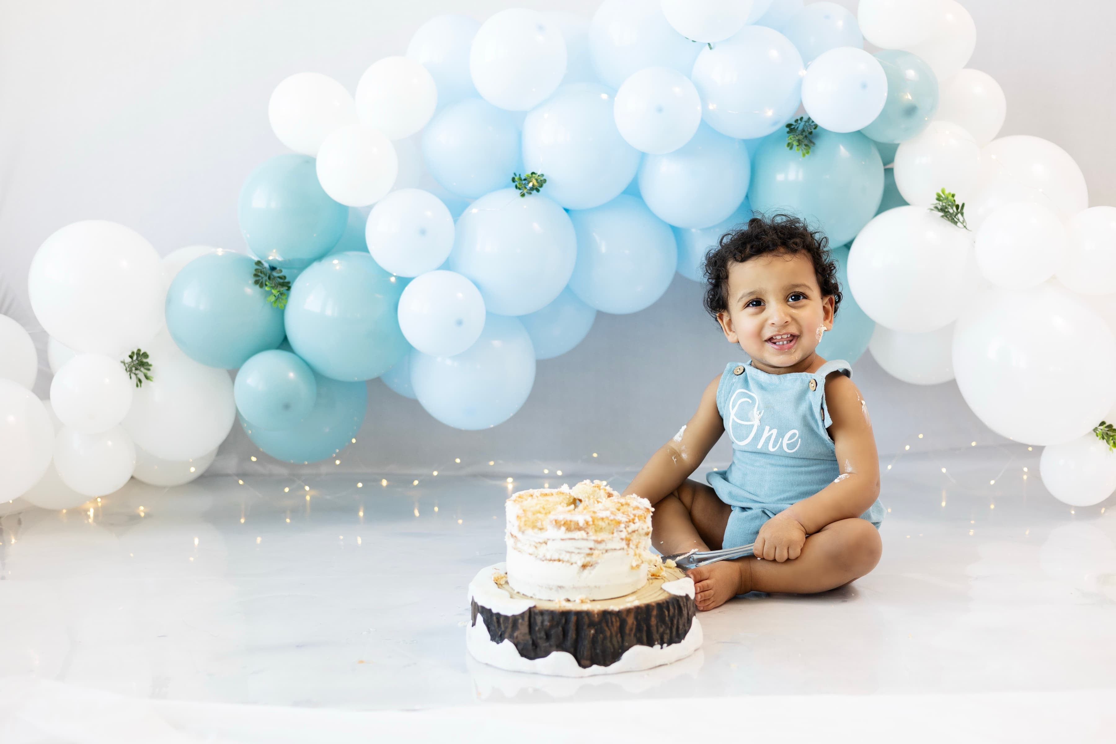 A one-year-old boy in a blue "One" romper sits beside his birthday cake, smiling directly at the camera with frosting on his cheek, framed by a pastel balloon arch.